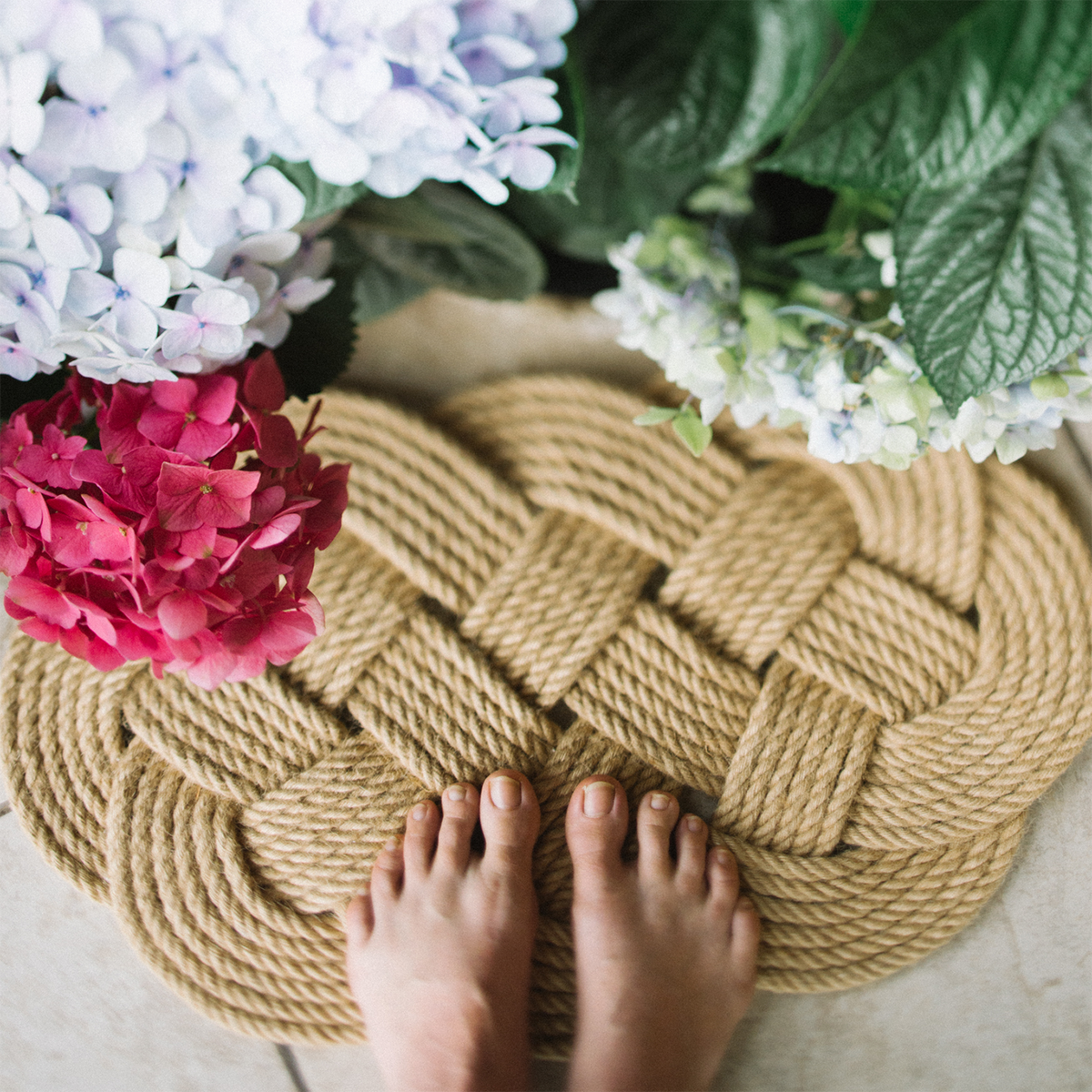 Bare feet on woven rope doormat surrounded by colorful hydrangea flowers, top view.