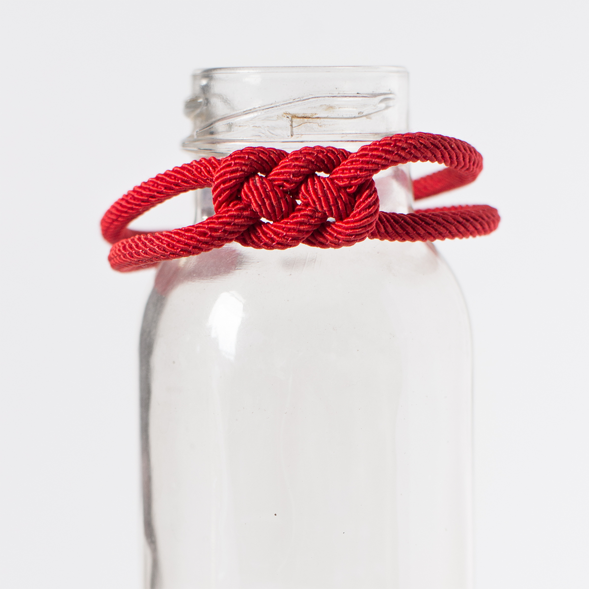 Red braided cord bracelet displayed on a glass bottle, minimalist white background